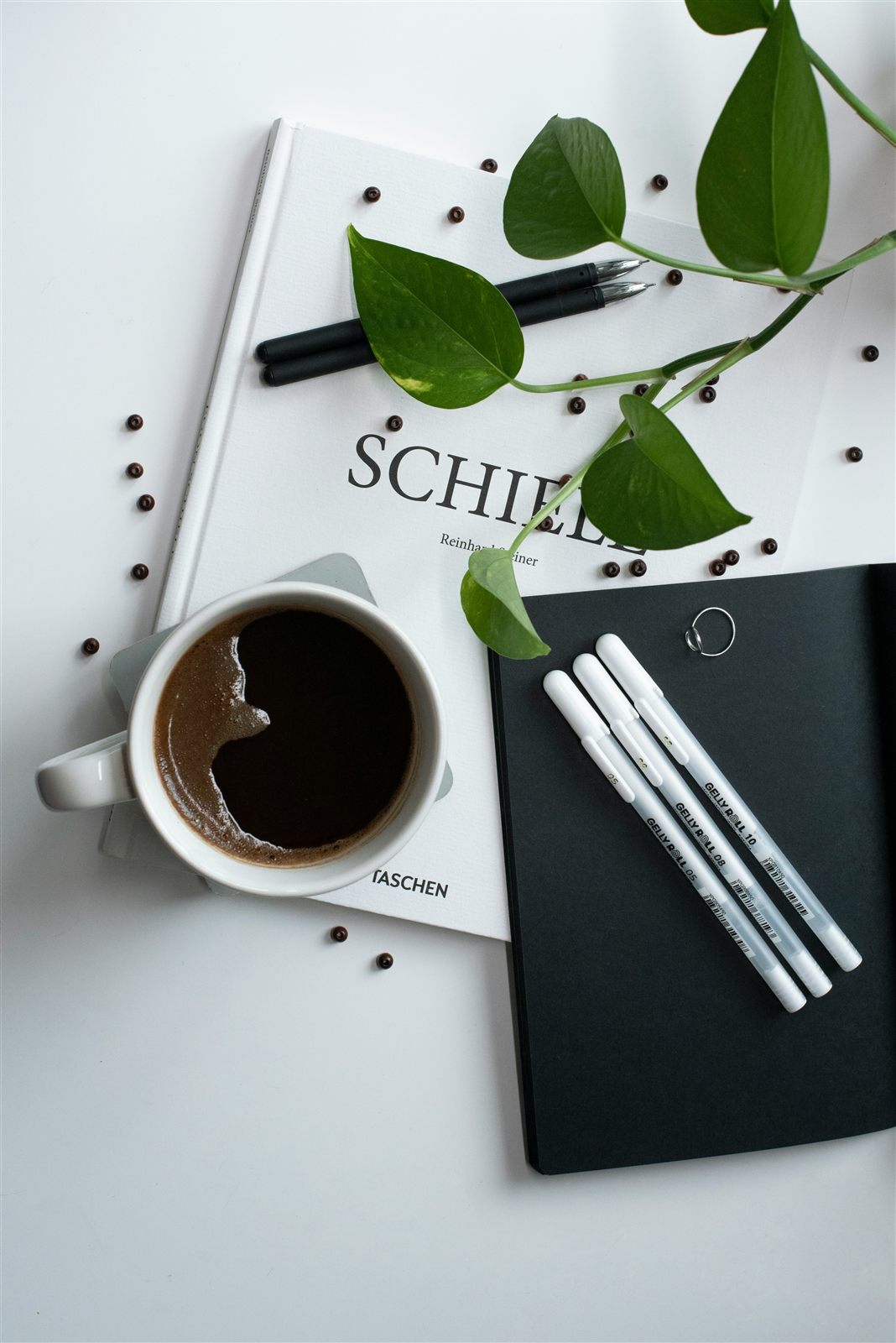 Photo of a cup of coffee, pens, and a small green plant on a desk beside an open notebook and a book