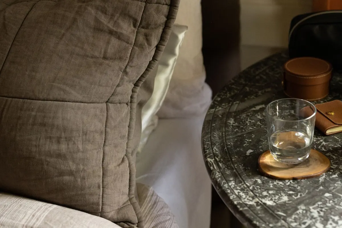 A glass of water on a round stone bedside table next to a linen pillow in soft evening light